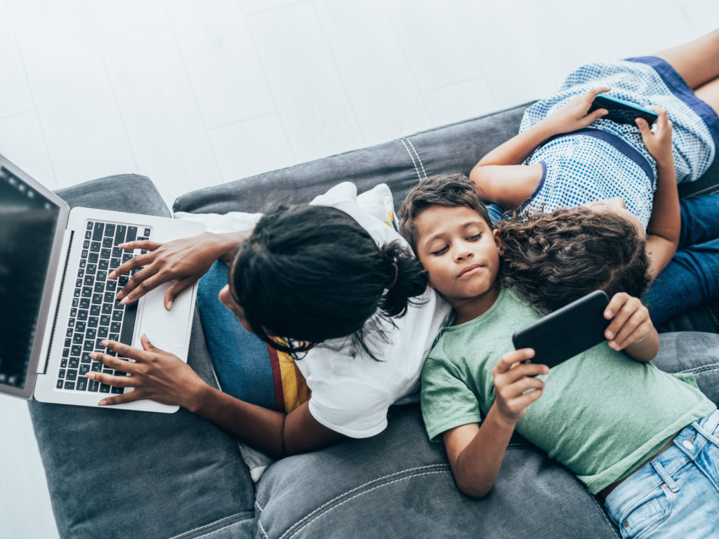 Siblings at home on their couch with various devices, including laptop and smartphone