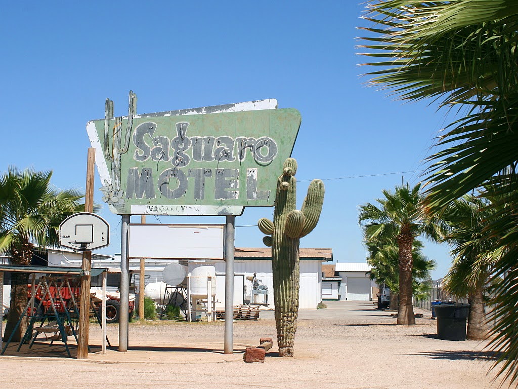 Distressed sign for Saguaro Motel in Aguila sirrounded by blue skies, palm trees, and a large cactus