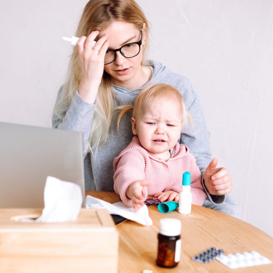 Portrait of young mother woman sitting at table near laptop, different pills blisters, with little baby girl, reading prescription for using spray medicine at home. Toddler crying, asking for bottle.